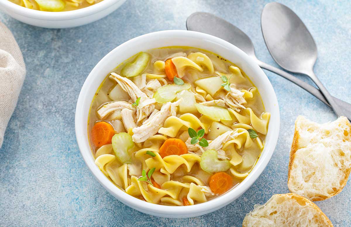 a bowl of rotisserie chicken noodle soup with chicken noodles and carrots. some bread on side