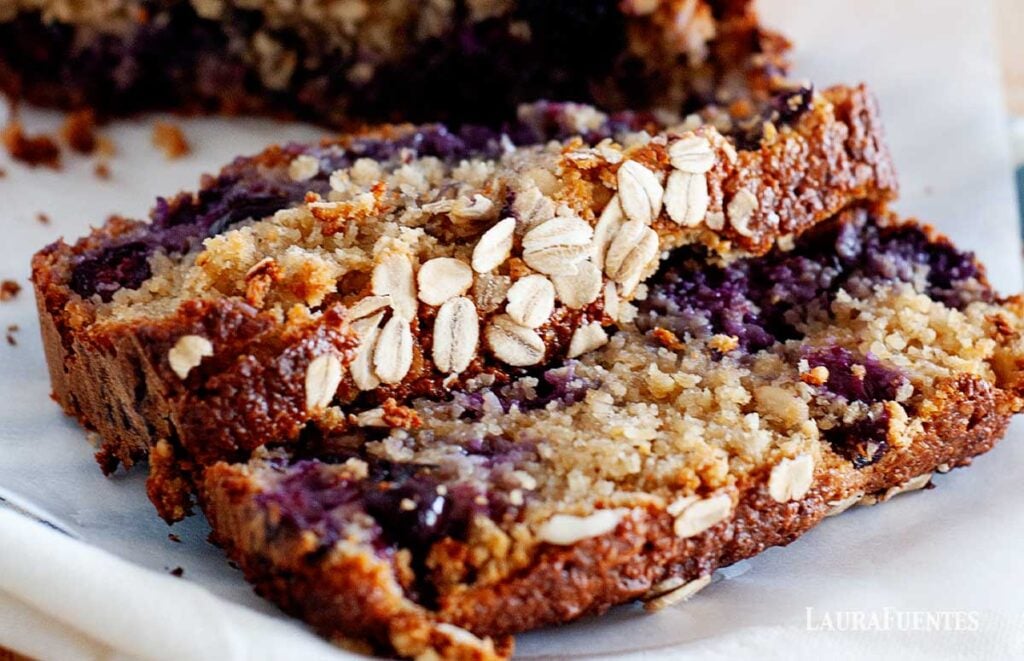 image: closeup view of breakfast bread loaf with blueberries and topped with oats. Two slices cut in front of the loaf.