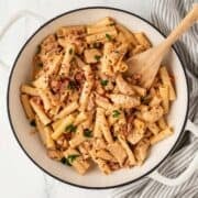 overhead view of a skillet filled with chicken pasta with a creamy sun dried tomato sauce a wooden spoon resting inside