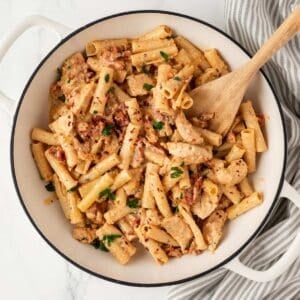 overhead view of a skillet filled with chicken pasta with a creamy sun dried tomato sauce a wooden spoon resting inside