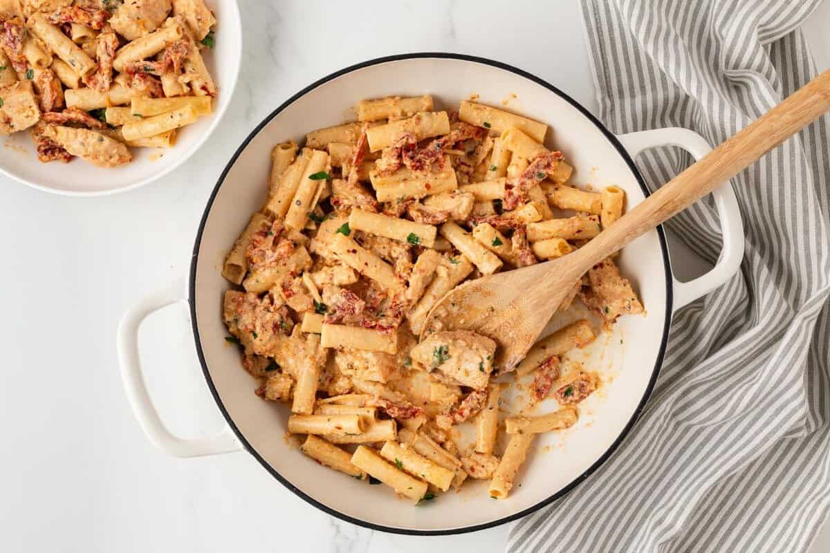 overhead view of a skillet filled with chicken pasta with a creamy sun dried tomato sauce a wooden spoon resting inside, and a bowl in the corner filled with some.