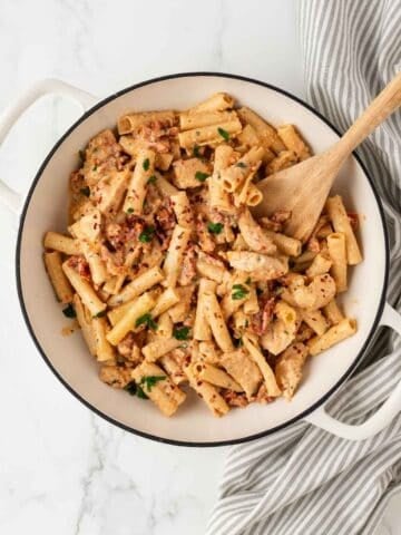 overhead view of a skillet filled with chicken pasta with a creamy sun dried tomato sauce a wooden spoon resting inside