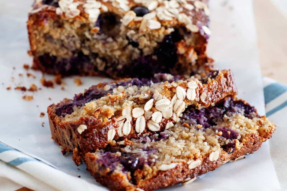 closeup view of breakfast bread loaf with blueberries and topped with oats; two slices cut in front of the loaf
