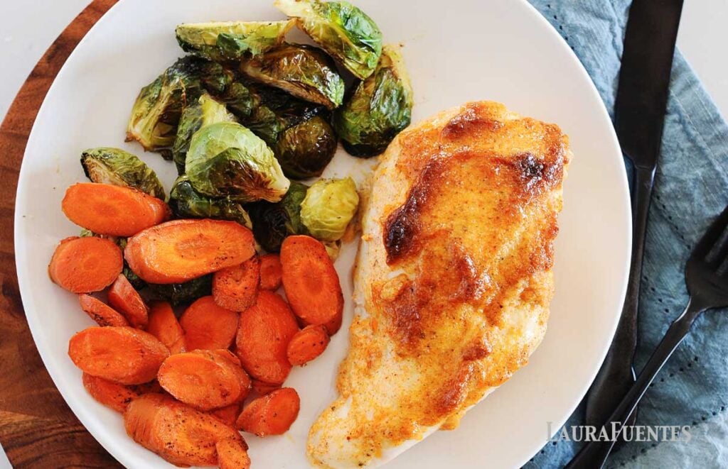 juicy baked chicken breast on a white plate with roasted brussel sprout halves and carrot slices.
