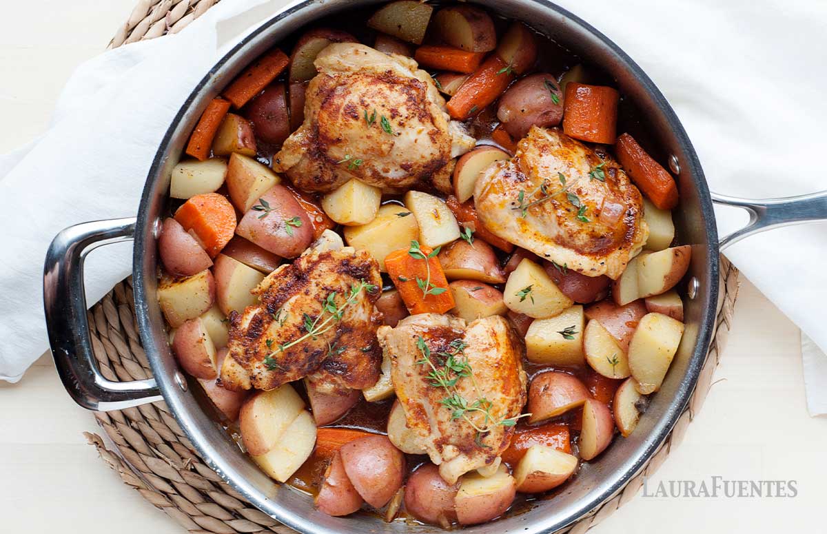 image: large skillet filled with cooked potato chunks, carrot chunks and chicken thighs. 