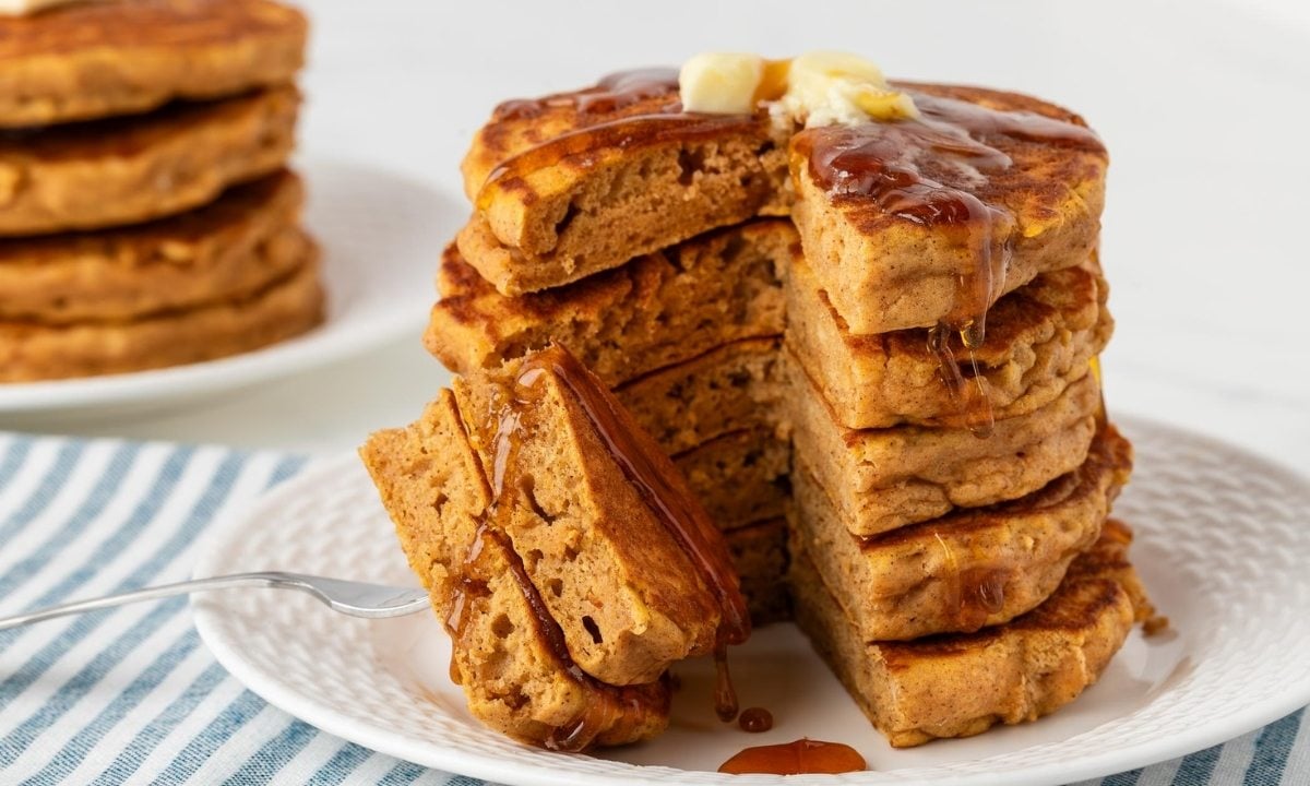 stack of fluffy sweet potato pancakes on a white plate topped with butter and pancake syrup. A piece of pancake has been sliced to show fluffy texture