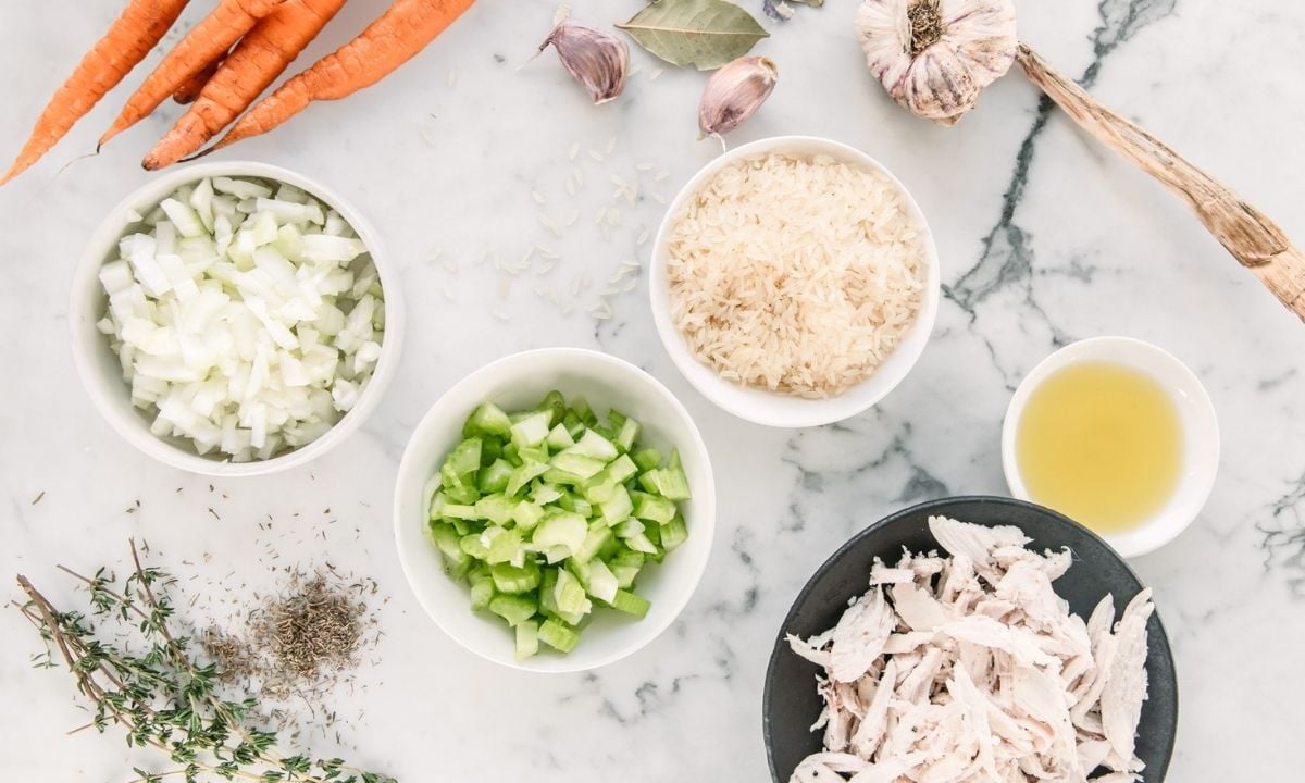 Ingredients for chicken and rice soup, including shredded chicken, white rice, onion, celery, carrots, garlic, chicken broth, olive oil, thyme, and bay leaves arranged on a marble countertop.