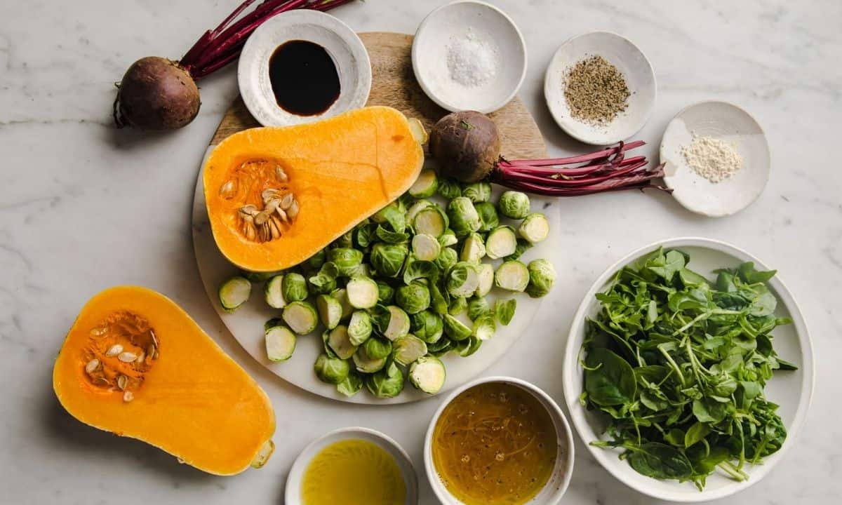 Ingredients for roasted fall vegetables, including butternut squash, Brussels sprouts, beets, leafy greens, and seasonings.