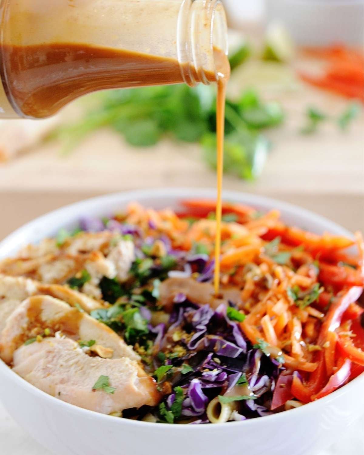 dressing being poured over a bowl of thai chicken pasta salad with carrots, bell peppers and cabbage
