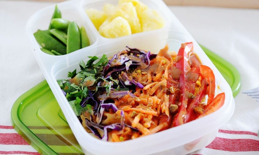 a lunchbox with thai chicken pasta salad in the biggest compartment, fruit and veggies in the smallest ones.
