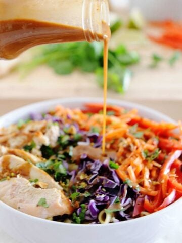 dressing being poured over a bowl of thai chicken pasta salad with carrots, bell peppers and cabbage