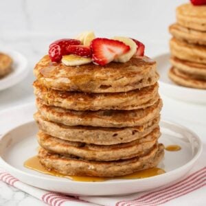 stack of whole wheat pancakes on a plate topped with sliced strawberries and banana with syrup on a plate.