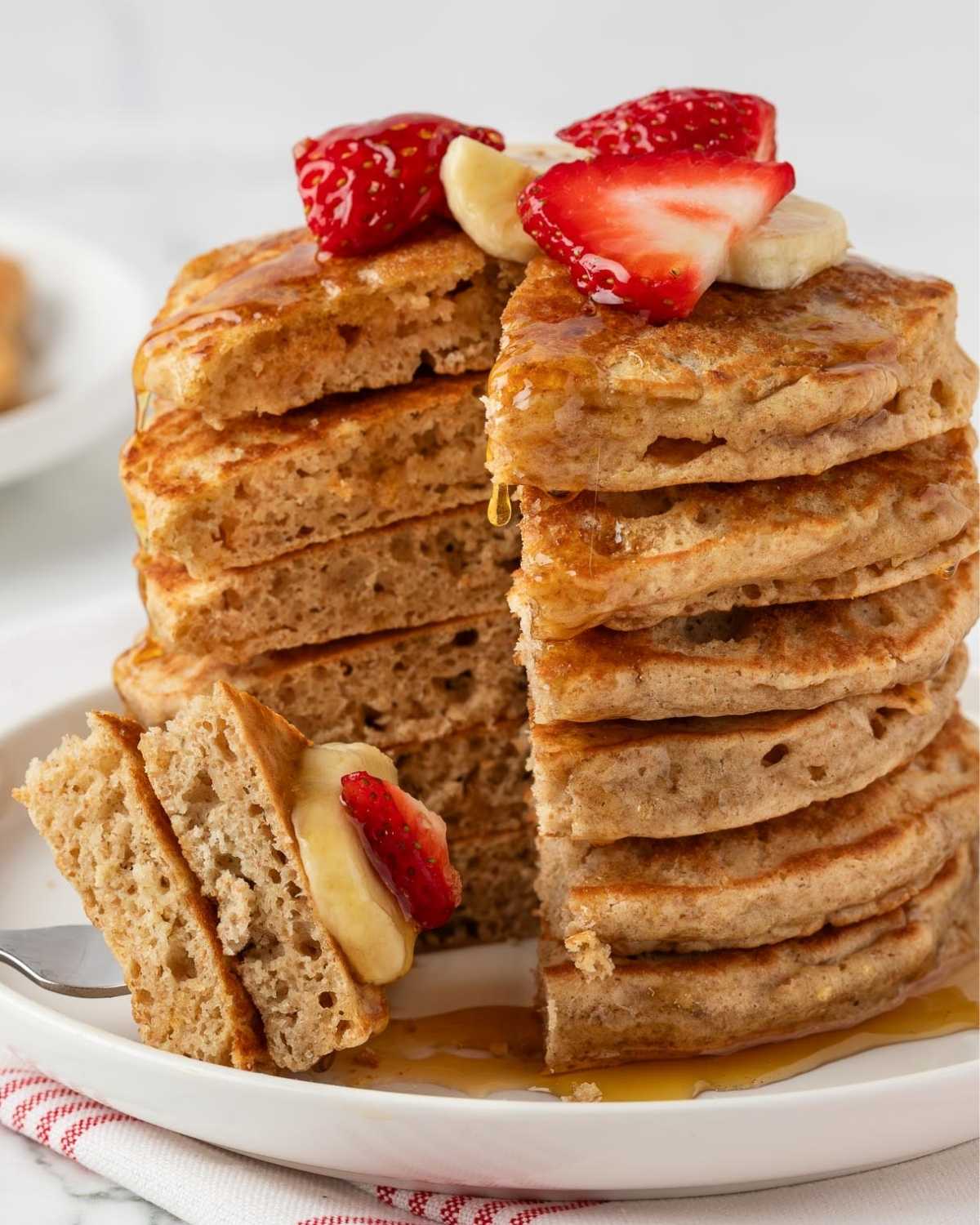 stack of whole wheat pancakes on a plate topped with sliced strawberries and banana with syrup on a plate. A slice of whole wheat pancakes on a fork showing fluffy texture of recipe