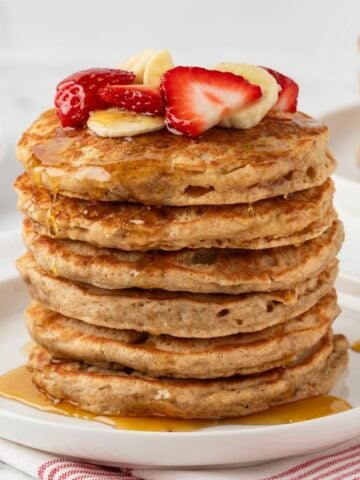 stack of whole wheat pancakes on a plate topped with sliced strawberries and banana with syrup on a plate.
