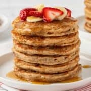 stack of whole wheat pancakes on a plate topped with sliced strawberries and banana with syrup on a plate