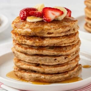 stack of whole wheat pancakes on a plate topped with sliced strawberries and banana with syrup on a plate