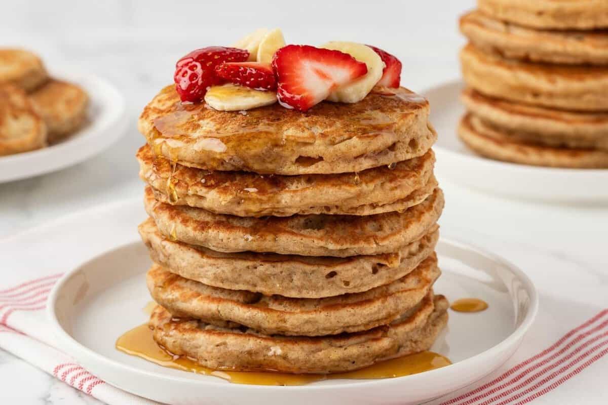 stack of whole wheat pancakes on a plate topped with sliced strawberries and banana with syrup on a plate