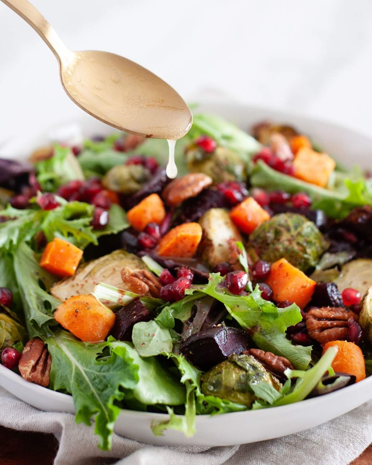 a spoon drizzling dressing on a large salad bowl with roasted winter veggies and greens