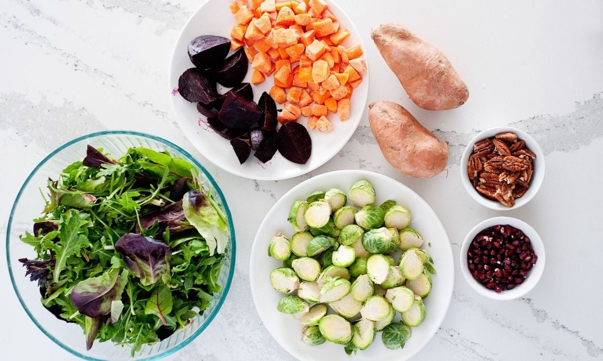 bowls with green, Brussels sprouts, cubed sweet potatoes and beets in a plate next to two sweet potatoes, and two small bowls with nuts