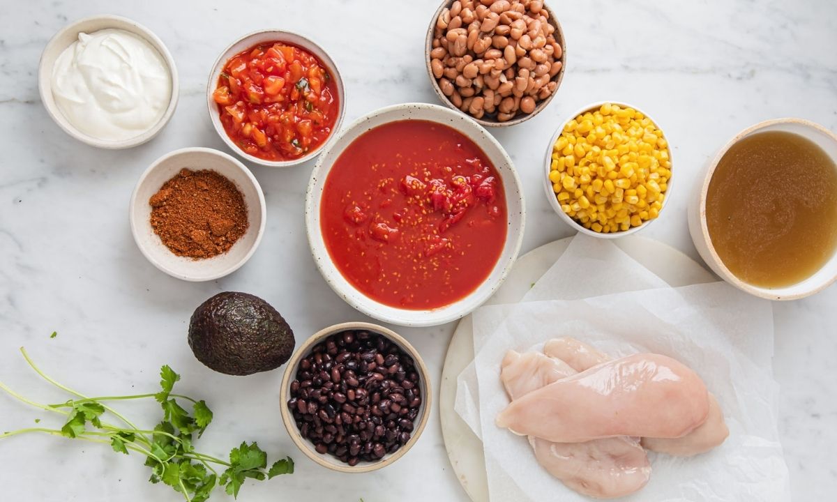 chicken taco soup ingredients with chicken breasts, diced tomatoes, black beans, pinto beans, corn, chicken broth, taco seasoning, salsa, avocado, sour cream, and cilantro.