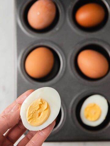 a hand holding half a boiled egg above a muffin tin of boiled eggs