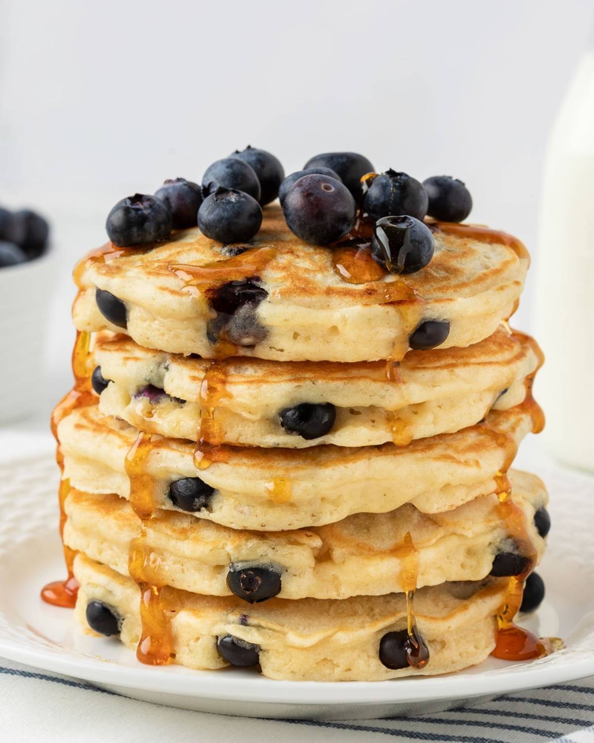 Fluffy stack of homemade blueberry pancakes on a plate. They're topped with more blueberries and a light drizzle of pancake syrup.