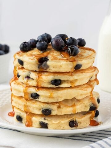Fluffy stack of homemade blueberry pancakes on a plate. They're topped with more blueberries and a light drizzle of pancake syrup.