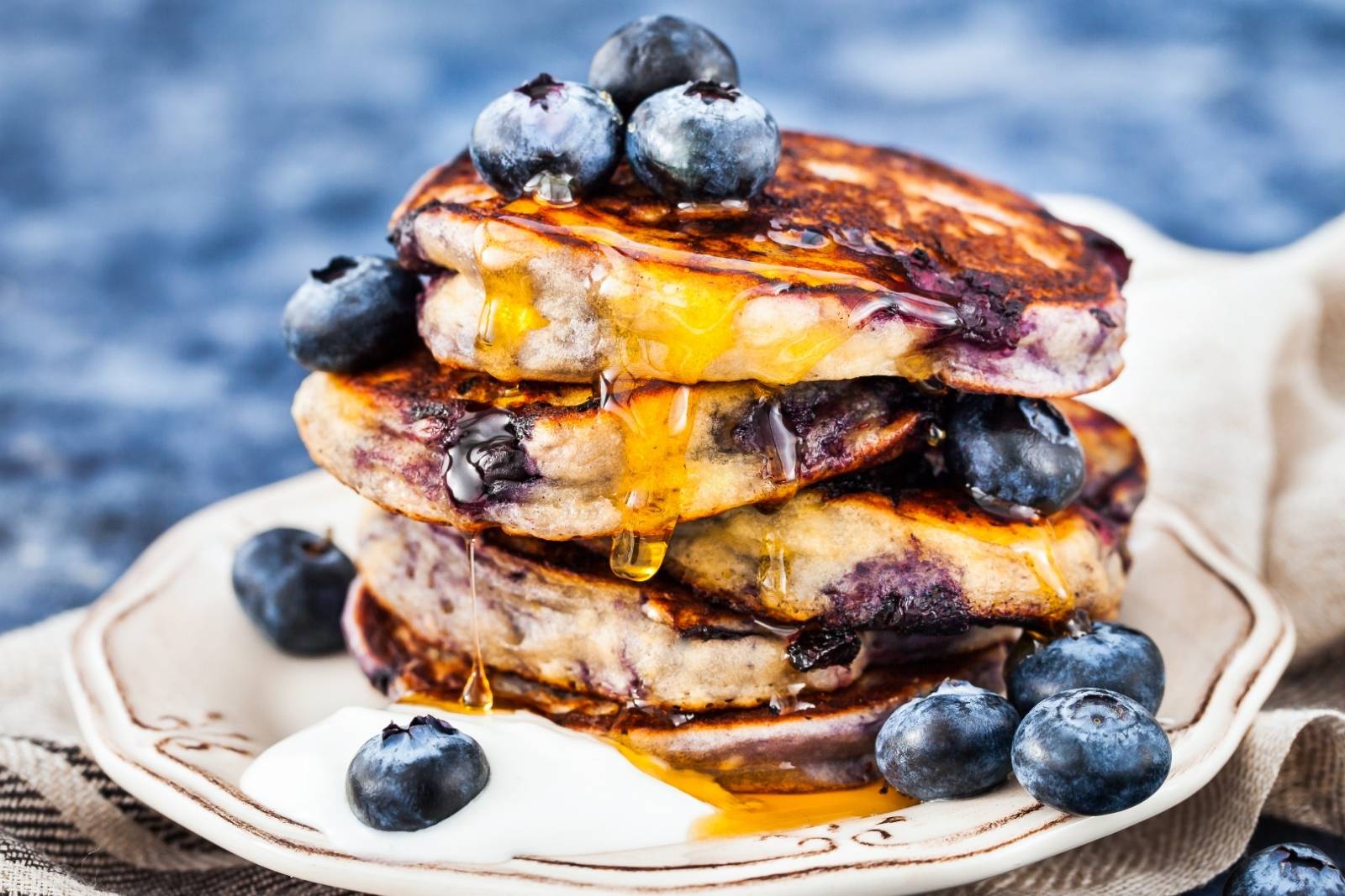 stack of blueberry pancakes bursting with juicy blueberries inside. topped with a few blueberries and syrup, with blueberries and a dollop of yogurt on a plate