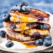 stack of blueberry pancakes bursting with juicy blueberries inside. topped with a few blueberries and syrup, with blueberries and a dollop of yogurt on a plate