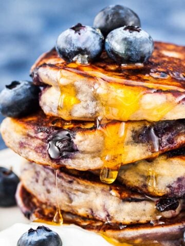 stack of blueberry pancakes bursting with juicy blueberries inside. topped with a few blueberries and syrup, with blueberries and a dollop of yogurt on a plate