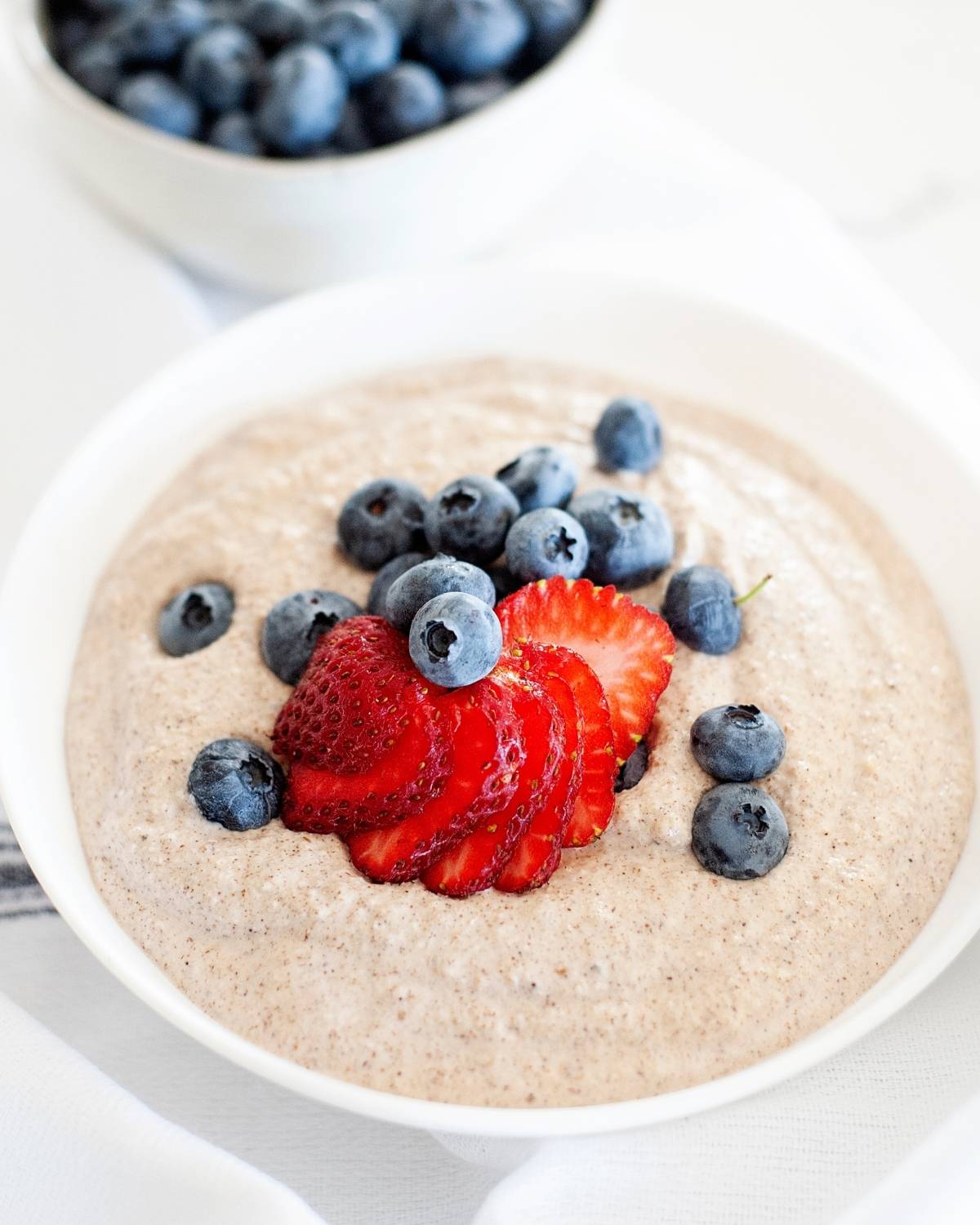 bowl of paleo oatmeal with berries on top, and a small bowl of bluberries behind