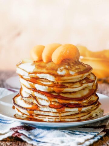 Fluffy stack of peach pancakes topped with peaches and pancake syrup on a plate