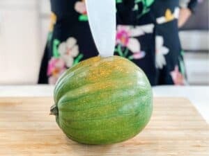 a whole acorn squash on a wooden board being poke with a knife