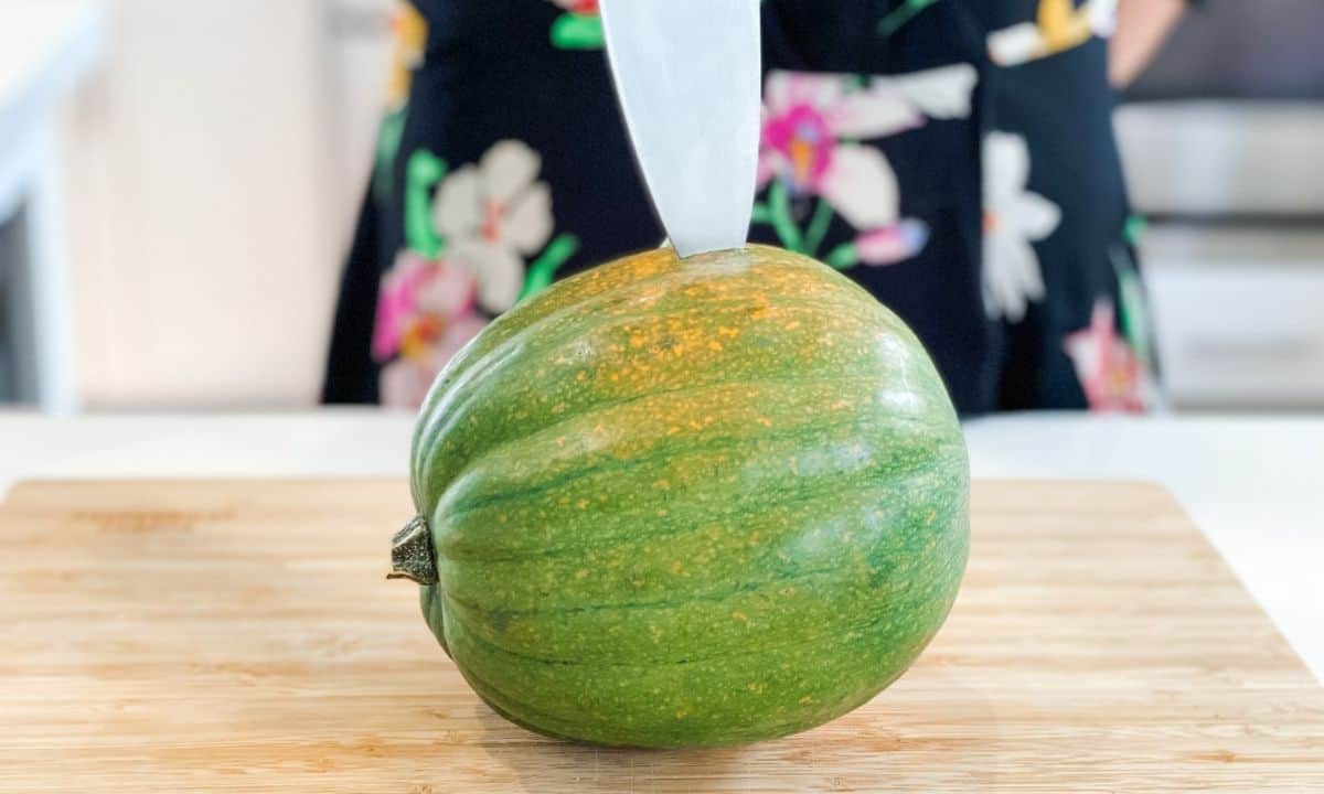a whole acorn squash on a wooden board being poke with a knife