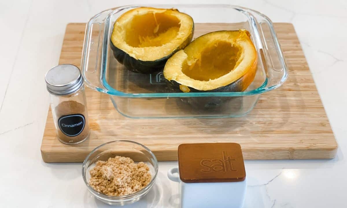 two cooked acorn squash halves on a microwave dish, a bowl with brown sugar, a jar of cinnamon, and salt