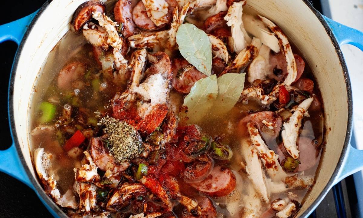 Chicken gumbo simmering in a Dutch oven with shredded chicken, sliced sausage, bell peppers, celery, bay leaves, broth, and seasonings.
