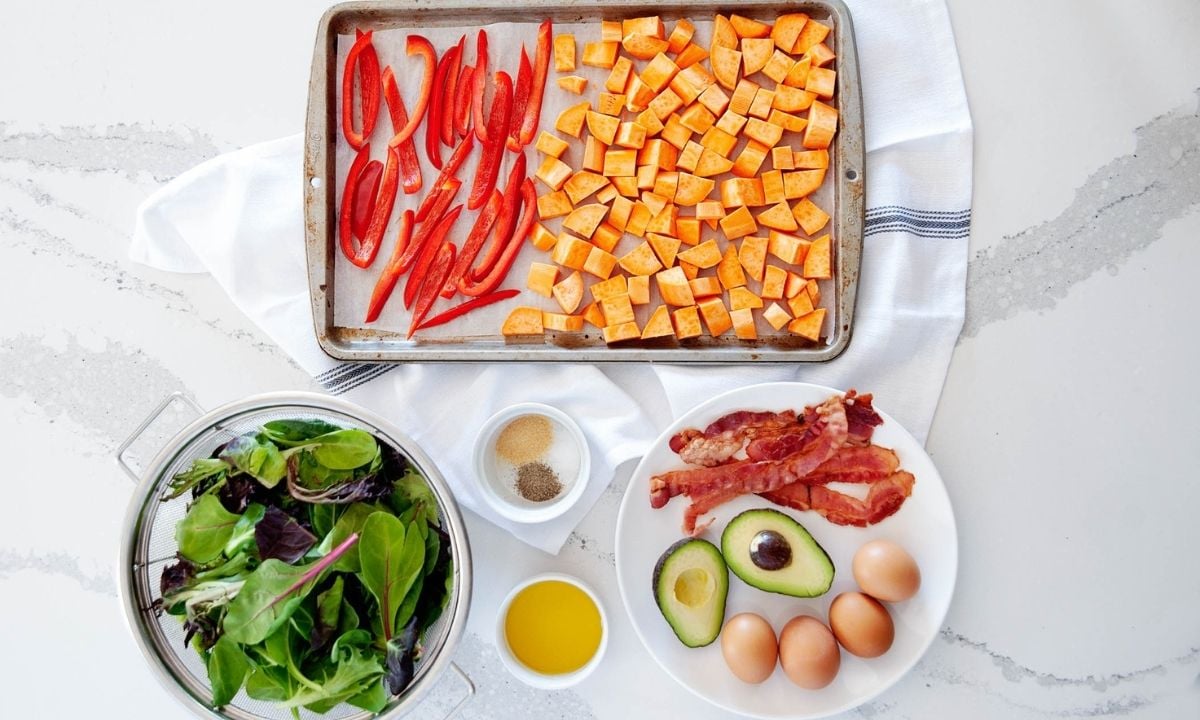 ingredients for breakfast salad: baking sheet with bell pepper and sweet potato cubes, a bowl of greens, seasonings, oil, avocado, eggs, and cooked bacon