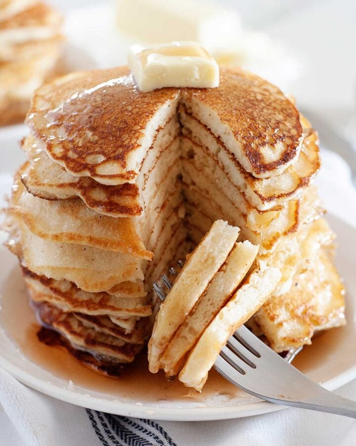 stack of old fashioned pancakes made with buttermilk at home topped with butter and drizzled with maple syrup that has been sliced to show the inside fluffy texture