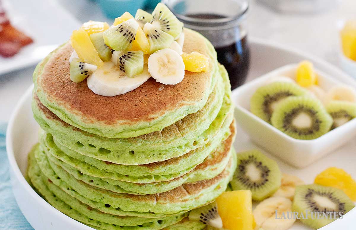 large stack of green pancakes with sliced fruit and syrup to one side