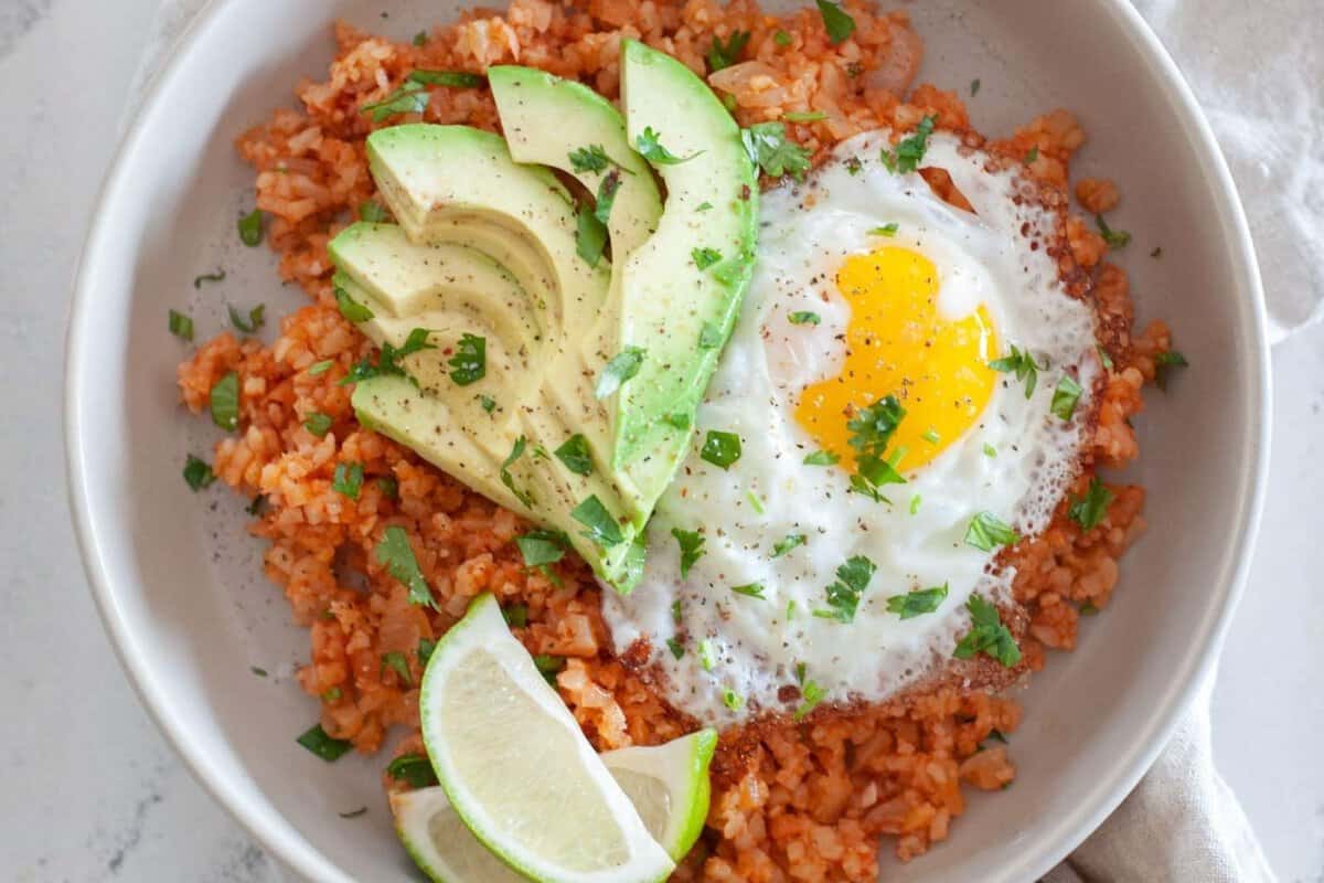 plate of spanish cauliflower rice, topped with fried egg sliced avocado, and limes 
