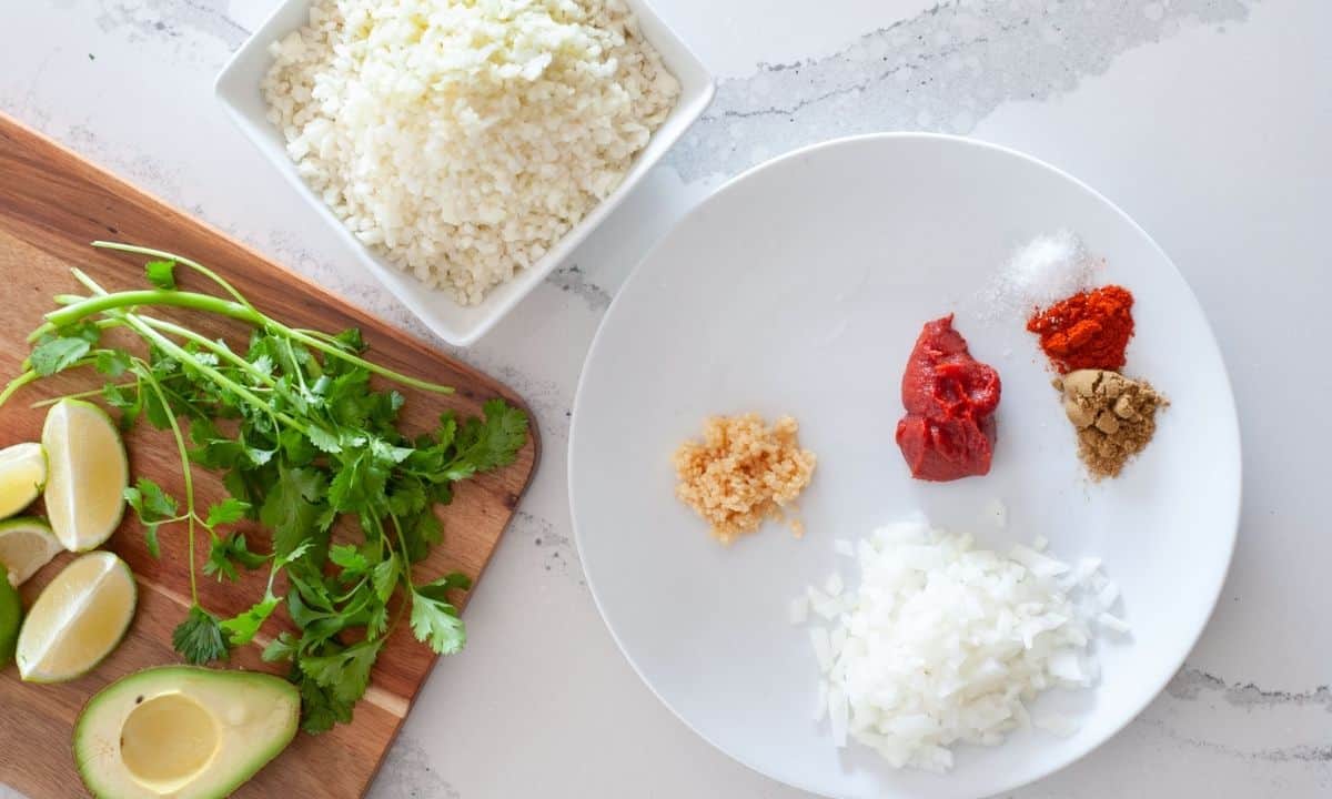 ingredients for Spanish cauliflower rice: cauliflower rice, olive oil, onions, garlic, cumin, paprika, salt, tomato paste, lime wedges, and fresh cilantro