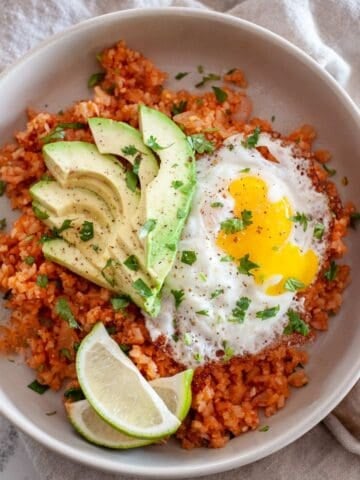 plate of spanish cauliflower rice, topped with fried egg sliced avocado, and limes