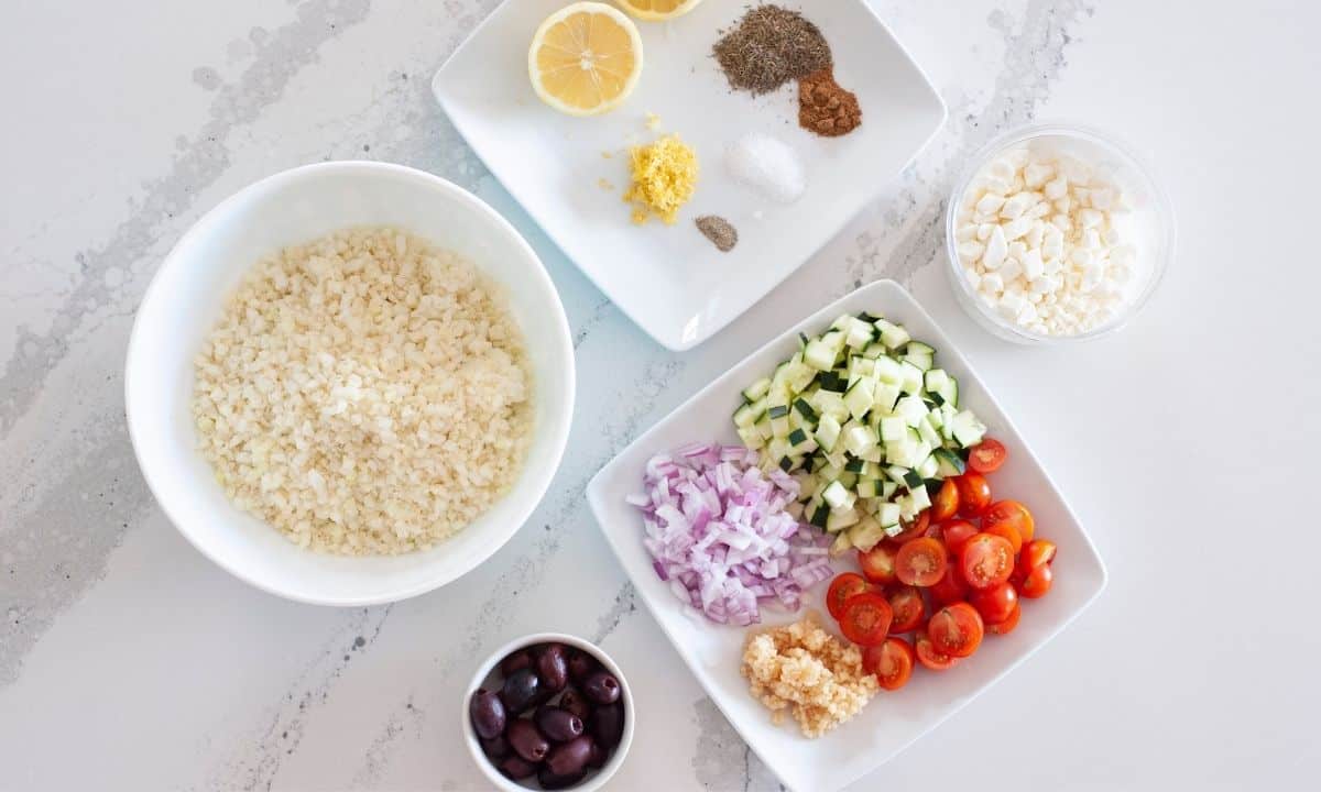 Ingredients for Mediterranean cauliflower rice, including cauliflower rice, tomatoes, cucumber, onion, garlic, olives, lemon, feta cheese, and seasonings.