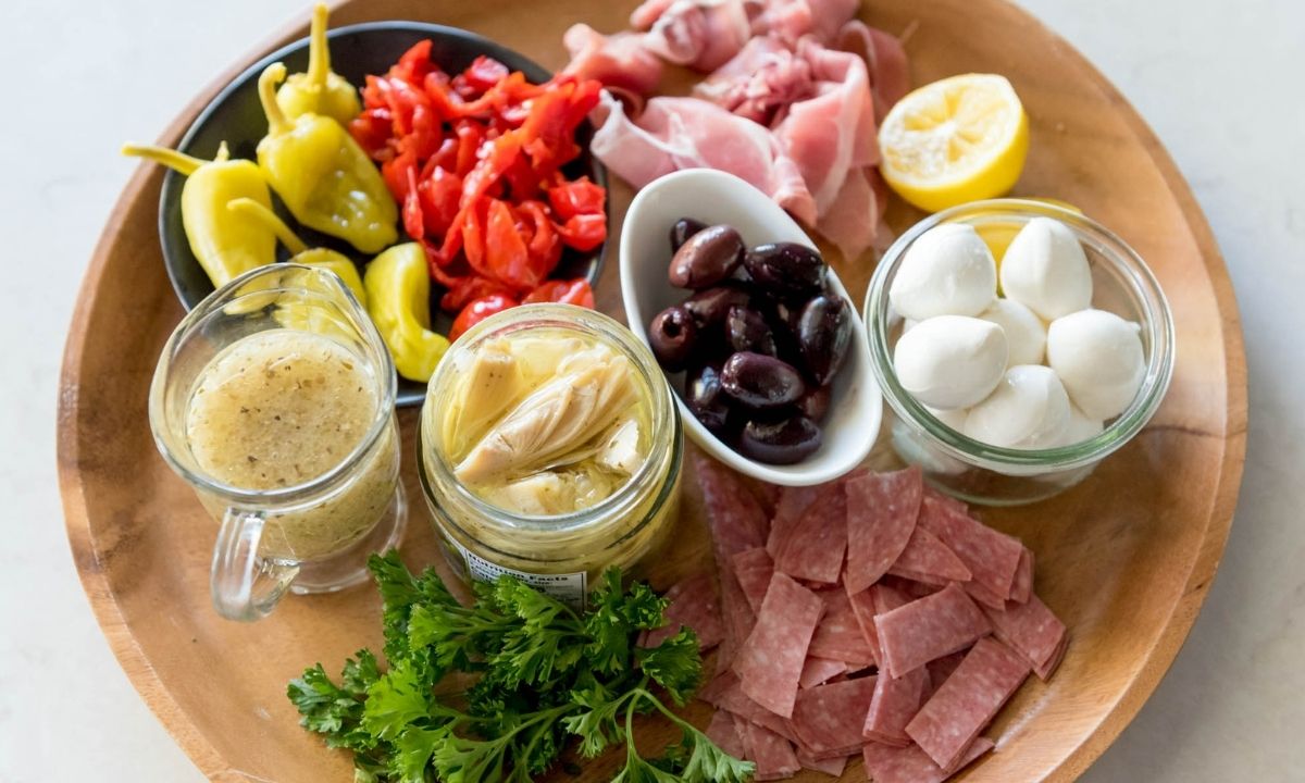 a board with olives, salami, pepperoncini, prosciutto, peppers, lemon, and dressing