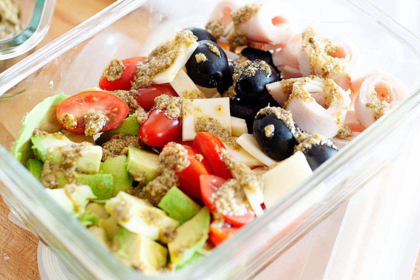 pesto salad with avocado, tomatoes, olives, ham and cheese in a glass lunch container