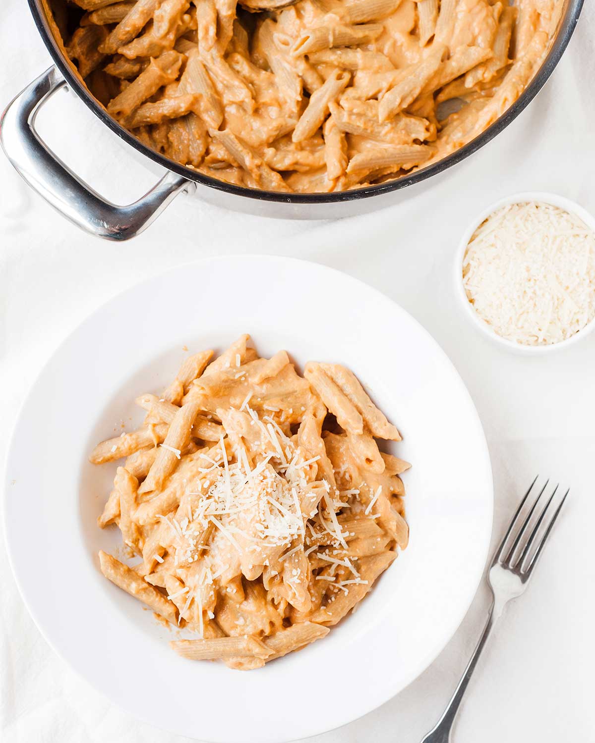 overhead view of plate filled with penne pasta tossed in a creamy pumpkin sauce topped with some parmesan cheese and showing the rest of the recipe inside a skillet (not served)