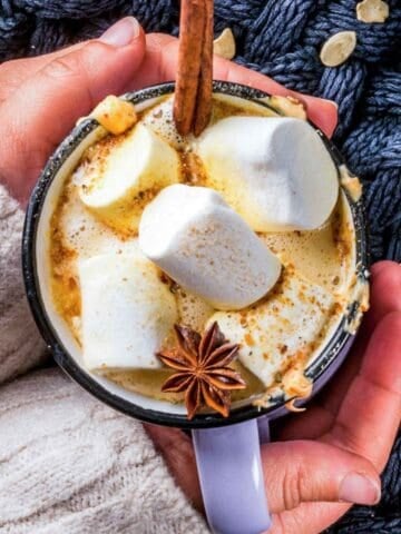 two hands holding a mug of pumpkin hot chocolate topped with marshmallows and a cinnamon stick