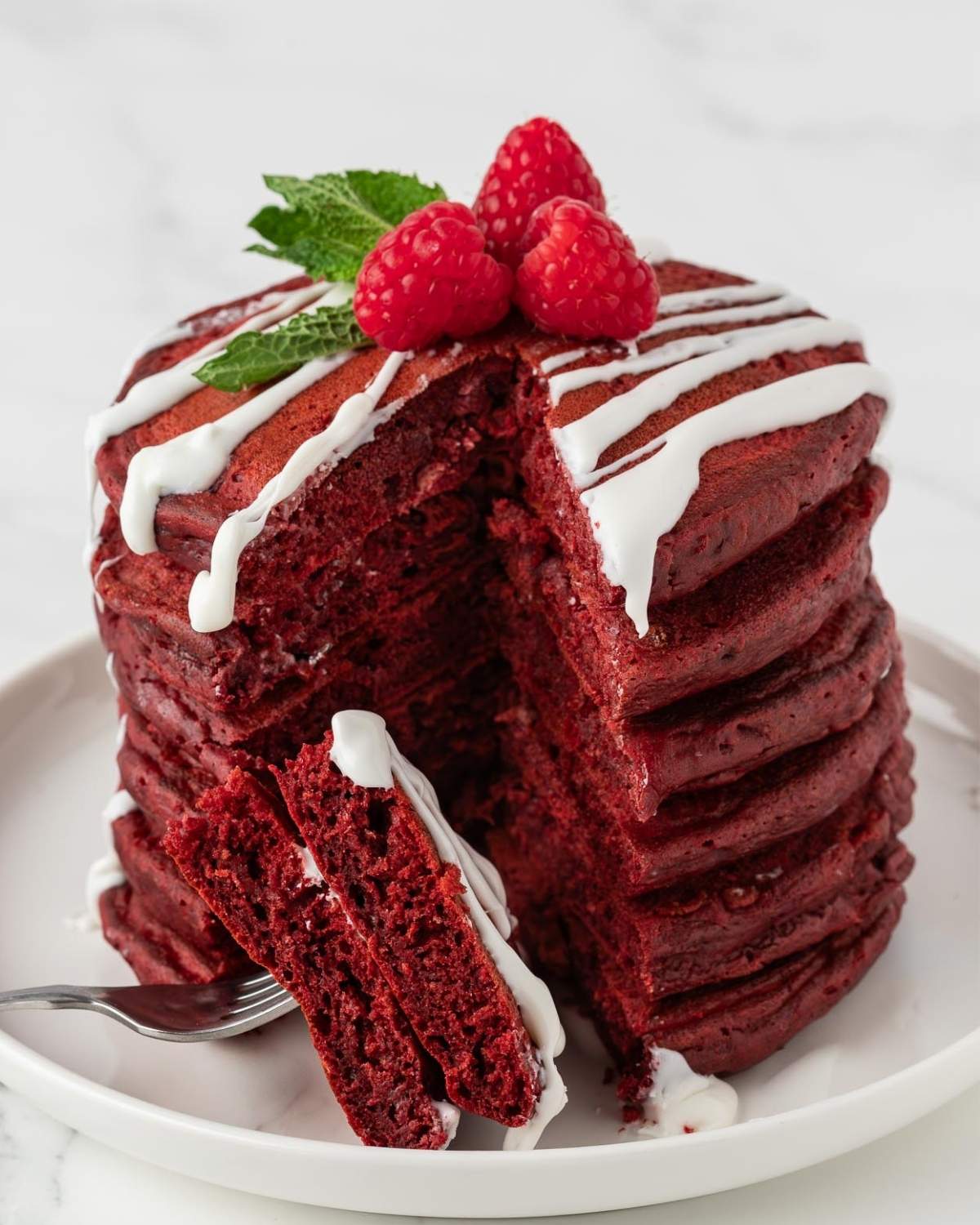stack of fluffy red velvet pancakes on a plate that's been sliced to show the rich color and texture topped with a light drizzle of white sugar glaze topped with raspberries