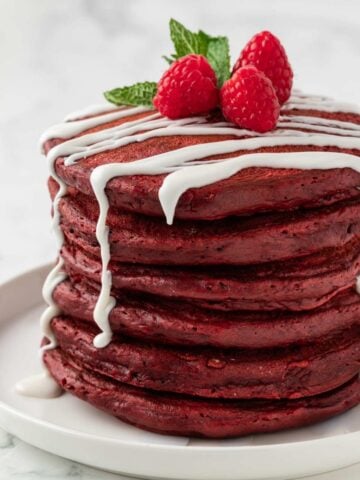 stack of fluffy red velvet pancakes on a plate with a light drizzle of white sugar glaze topped with raspberries