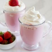 two clear mugs of pink hot chocolate topped with whipped cream next to a small dish of strawberries